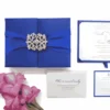 A royal blue wedding invitation suite displayed as a flat lay on a white background. On the left, a square gatefold box in blue silk is wrapped with a matching satin ribbon and secured by a large, ornate silver filigree brooch. To the right, the open invitation shows elegant script and blue corner tabs. Below the box are a matching RSVP card, an envelope printed with the names "Oliver and Emily," and a cluster of vibrant pink gladiolus flowers in the bottom left corner. The composition is brightly lit, highlighting the rich blue fabric and delicate stationery details.