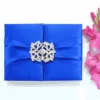 A rectangular, royal blue silk gatefold invitation box centered on a white background. A wide, matching blue satin ribbon wraps horizontally around the box, gathered in the center by a large, ornate silver brooch with a complex, lace-like filigree design. To the right of the box, a cluster of vibrant pink gladiolus flowers adds a pop of contrasting color. The flat lay composition is brightly lit, emphasizing the smooth sheen of the satin and the rich texture of the silk fabric.