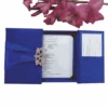 A royal blue wedding invitation suite displayed as a flat lay on a white background. On the left, a square gatefold box in blue silk is wrapped with a matching satin ribbon and secured by a large, ornate silver filigree brooch. To the right, the open invitation shows elegant script and blue corner tabs. Below the box are a matching RSVP card, an envelope printed with the names "Oliver and Emily," and a cluster of vibrant pink gladiolus flowers in the bottom left corner. The composition is brightly lit, highlighting the rich blue fabric and delicate stationery details.