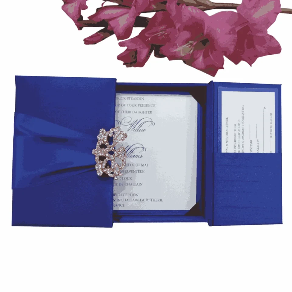 A royal blue wedding invitation suite displayed as a flat lay on a white background. On the left, a square gatefold box in blue silk is wrapped with a matching satin ribbon and secured by a large, ornate silver filigree brooch. To the right, the open invitation shows elegant script and blue corner tabs. Below the box are a matching RSVP card, an envelope printed with the names "Oliver and Emily," and a cluster of vibrant pink gladiolus flowers in the bottom left corner. The composition is brightly lit, highlighting the rich blue fabric and delicate stationery details.