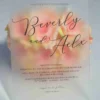 A Frosted acrylic wedding invitation is displayed at an angle, leaning against a large bouquet of soft peach and light pink roses. The card features elegant Black foil text and minimalist graphics.