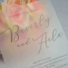 A close-up, angled shot of a frosted acrylic wedding invitation placed in front of a bouquet of peach and pink roses. The frosted finish of the acrylic softly blurs the floral colors behind it, creating a dreamy, watercolor effect.
