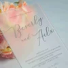 A close-up, angled shot of a frosted acrylic wedding invitation placed in front of a bouquet of peach and pink roses. The frosted finish of the acrylic softly blurs the floral colors behind it, creating a dreamy, watercolor effect.