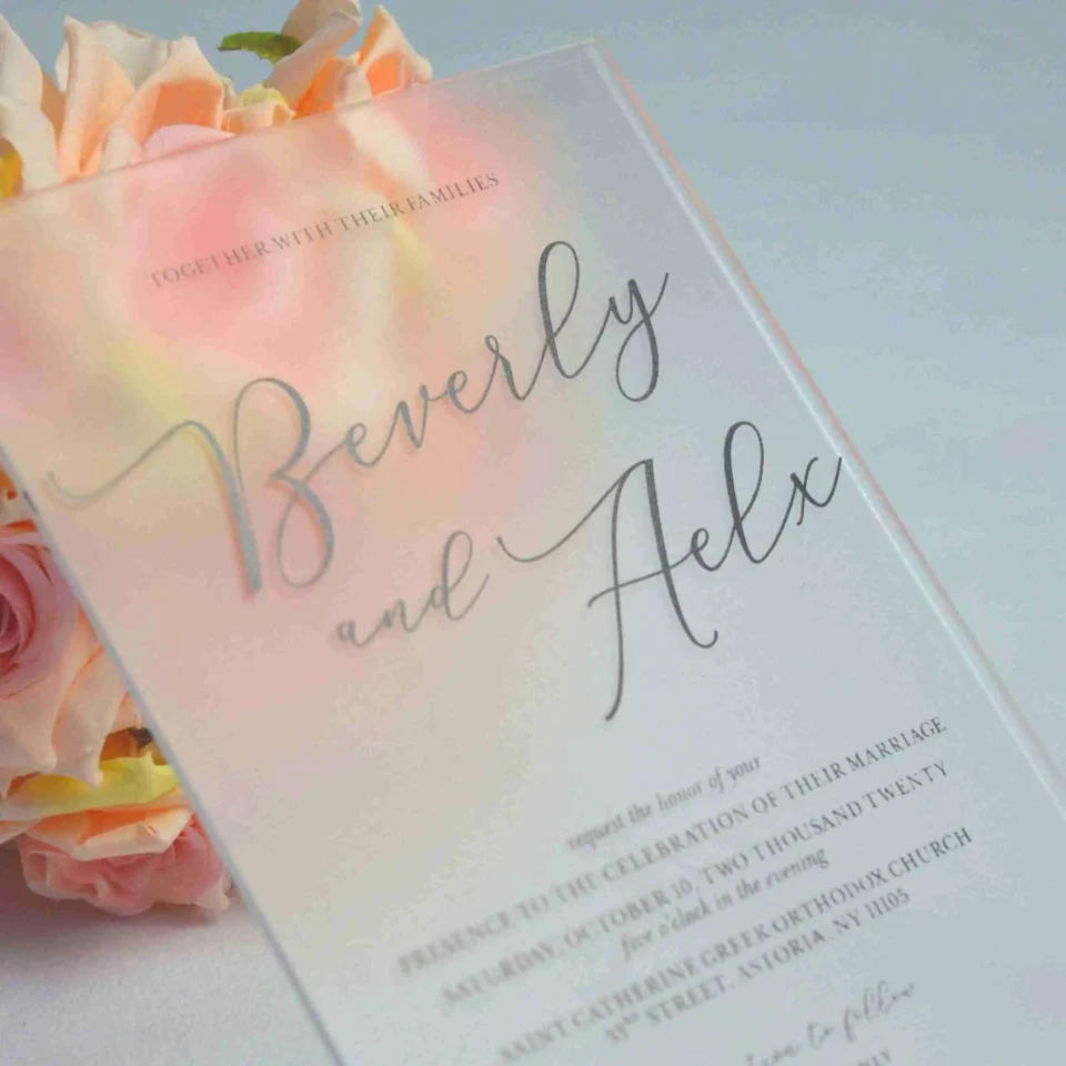 A close-up, angled shot of a frosted acrylic wedding invitation placed in front of a bouquet of peach and pink roses. The frosted finish of the acrylic softly blurs the floral colors behind it, creating a dreamy, watercolor effect.