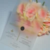 An elegant Frosted acrylic wedding invitation is shown leaning against a lush bouquet of soft peach and pink roses on a white surface. The invitation features a minimalist design with gold foil text and a delicate gold geometric line pattern at the bottom. The text on the clear card includes Details