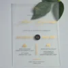 A frosted acrylic wedding invitation is laid flat on a light gray surface with a green leaf partially visible behind the top right corner. The invitation features a modern layout with a mix of gold foil and black text.