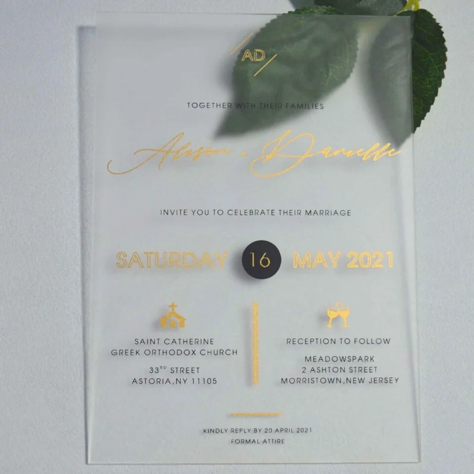 A frosted acrylic wedding invitation is laid flat on a light gray surface with a green leaf partially visible behind the top right corner. The invitation features a modern layout with a mix of gold foil and black text.