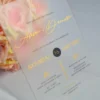 An elegant Frosted acrylic wedding invitation is shown leaning against a lush bouquet of soft peach and pink roses on a white surface. The invitation features a minimalist design with gold foil text and a delicate gold geometric line pattern at the bottom. The text on the clear card includes Details