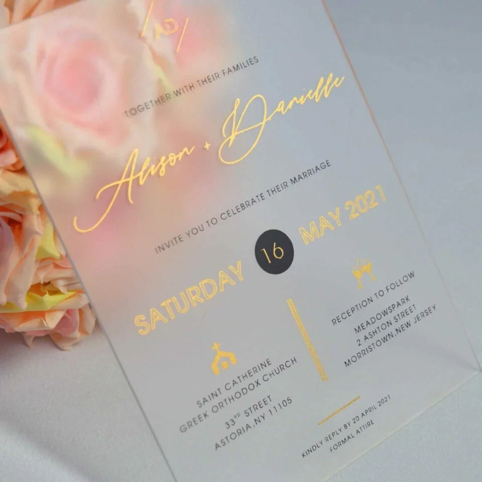 An elegant Frosted acrylic wedding invitation is shown leaning against a lush bouquet of soft peach and pink roses on a white surface. The invitation features a minimalist design with gold foil text and a delicate gold geometric line pattern at the bottom. The text on the clear card includes Details