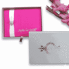 A rectangular, vibrant hot pink silk wedding invitation folio is centered against a white background. A wide, shimmering white or silver satin ribbon runs vertically down the left side, cinched at the middle by a round, sparkling rhinestone button brooch. In the bottom right corner, the names "Mathew and Michelle" are printed in an elegant, silver-toned cursive script. To the right of the folio, a tall stalk of bright pink gladiolus blossoms adds a soft, floral frame to the composition.