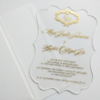 A transparent acrylic wedding invitation is shown partially tucked into a white paper pocket or sleeve. The image is shot at an angle on a white surface, highlighting the clarity of the invitation and the clean, minimalist presentation.