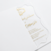 A transparent acrylic wedding invitation is shown partially tucked into a white paper pocket or sleeve. The image is shot at an angle on a white surface, highlighting the clarity of the invitation and the clean, minimalist presentation.