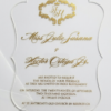 A transparent acrylic wedding invitation with a custom-contoured, wavy scalloped edge. The invitation is presented on a clean white background with a corner of a white envelope visible on the left.