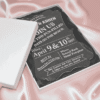 An angled, close-up view of an open white wedding invitation box resting on a soft, blush-toned fabric. The box features: A thick, clear acrylic invitation plaque** with a decorative scalloped edge, nestled inside on a black satin lining. Bold white typography on the acrylic card that reads "JOIN US FOR TWO FUN-FILLED DAYS ON THE BEACH" for a celebration on Location details for the "Round Rock Hotel Pompton" and RSVP information are clearly displayed at the bottom. The white lid of the box is partially visible, leaning against the base to reveal the luxury contents inside. The high-contrast design between the clear acrylic, white text, and dark lining creates a modern and high-end aesthetic.