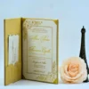 An open **gold velvet folio invitation** is displayed against a light blue background. The main invitation card on the right features an elegant gold metallic crest at the top, followed by traditional wedding or event text in sophisticated gold script. The left side of the folio includes a pocket holding coordinated **insert cards** for "Accommodations" and "Directions," also detailed with gold floral accents. To the right of the stationery sits a **pale peach rose** and a small **bronze Eiffel Tower** model, enhancing the luxurious and classic theme.