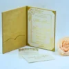 An open **gold velvet folio invitation** is shown from a high angle, lying flat against a light blue background. The main invitation card on the right features a regal gold crest and elegant gold script. In the foreground, a textured cream-colored envelope addressed to rests horizontally. A **pale peach rose** and a miniature **bronze Eiffel Tower** figurine are placed to the right, completing the luxurious and Parisian-inspired stationery suite.
