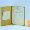 An open **gold velvet folio invitation** is displayed against a light blue background. The main invitation card on the right features an elegant gold metallic crest at the top, followed by traditional wedding or event text in sophisticated gold script. The left side of the folio includes a pocket holding coordinated **insert cards** for "Accommodations" and "Directions," also detailed with gold floral accents. To the right of the stationery sits a **pale peach rose** and a small **bronze Eiffel Tower** model, enhancing the luxurious and classic theme.