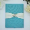 A top-down, centered shot of a **teal textured fabric invitation box** with a beach-themed design. The box features: * A wide, **Blue-colored silk sash** wrapped horizontally around the center. * A sparkling **crystal-encrusted starfish brooch** pinned at the center of the sash, serving as the main focal point. * A small teal pull-tab at the bottom edge for opening the box. * The corners of the frame are softly decorated with **white and pale yellow flowers**, along with a partial view of a bronze Eiffel Tower figurine in the upper right. * A clean, white background that makes the vibrant teal and shimmering brooch stand out.