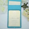 A top-down shot of an **open teal-colored invitation box** revealing a formal wedding invitation inside. The presentation includes: * A **Blue-colored invitation card** with a thin silver border, held in place by teal silk ribbon corners. * Elegant black script that reads "John & Julia" at the center, topped with a decorative silver crest. * The inner lid of the box features a **built-in pocket** containing a matching cream insert. * The scene is framed by **white roses** on the left and **pale yellow carnations** on the right, all set against a clean white background. * The soft lighting highlights the silk texture of the box and the crisp finish of the stationery.