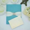 A top-down, centered shot of a **teal textured fabric invitation box** with a beach-themed design. The box features: * A wide, **Blue-colored silk sash** wrapped horizontally around the center. * A sparkling **crystal-encrusted starfish brooch** pinned at the center of the sash, serving as the main focal point. * A small teal pull-tab at the bottom edge for opening the box. * The corners of the frame are softly decorated with **white and pale yellow flowers**, along with a partial view of a bronze Eiffel Tower figurine in the upper right. * A clean, white background that makes the vibrant teal and shimmering brooch stand out.