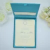 A top-down shot of an **open teal-colored invitation box** revealing a formal wedding invitation inside. The presentation includes: * A **Blue-colored invitation card** with a thin silver border, held in place by teal silk ribbon corners. * Elegant black script that reads "John & Julia" at the center, topped with a decorative silver crest. * The inner lid of the box features a **built-in pocket** containing a matching cream insert. * The scene is framed by **white roses** on the left and **pale yellow carnations** on the right, all set against a clean white background. * The soft lighting highlights the silk texture of the box and the crisp finish of the stationery.
