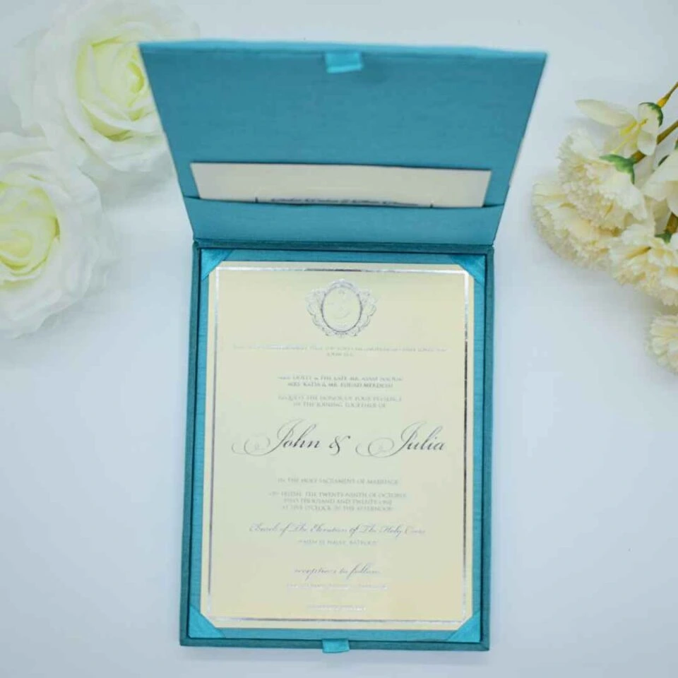 A top-down shot of an **open teal-colored invitation box** revealing a formal wedding invitation inside. The presentation includes: * A **Blue-colored invitation card** with a thin silver border, held in place by teal silk ribbon corners. * Elegant black script that reads "John & Julia" at the center, topped with a decorative silver crest. * The inner lid of the box features a **built-in pocket** containing a matching cream insert. * The scene is framed by **white roses** on the left and **pale yellow carnations** on the right, all set against a clean white background. * The soft lighting highlights the silk texture of the box and the crisp finish of the stationery.