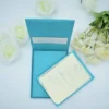 A top-down shot of an **open teal-colored invitation box** revealing a formal wedding invitation inside. The presentation includes: * A **Blue-colored invitation card** with a thin silver border, held in place by teal silk ribbon corners. * Elegant black script that reads "John & Julia" at the center, topped with a decorative silver crest. * The inner lid of the box features a **built-in pocket** containing a matching cream insert. * The scene is framed by **white roses** on the left and **pale yellow carnations** on the right, all set against a clean white background. * The soft lighting highlights the silk texture of the box and the crisp finish of the stationery.