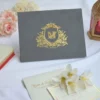 A landscape-oriented, grey velvet or suede invitation folder sits on a white surface, flanked by soft pink roses and a stack of small books. Centered on the cover is an intricate, regal gold foil crest featuring a crown, floral flourishes, and a central monogram. At the bottom of the crest, a gold ribbon banner displays the names along with the date.
