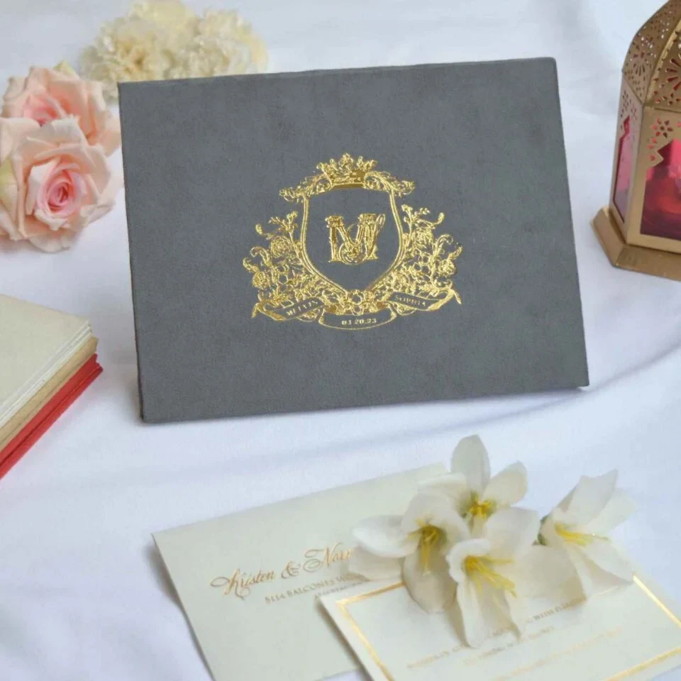 A landscape-oriented, grey velvet or suede invitation folder sits on a white surface, flanked by soft pink roses and a stack of small books. Centered on the cover is an intricate, regal gold foil crest featuring a crown, floral flourishes, and a central monogram. At the bottom of the crest, a gold ribbon banner displays the names along with the date.