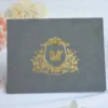 A landscape-oriented, grey velvet or suede invitation folder sits on a white surface, flanked by soft pink roses and a stack of small books. Centered on the cover is an intricate, regal gold foil crest featuring a crown, floral flourishes, and a central monogram. At the bottom of the crest, a gold ribbon banner displays the names along with the date.