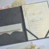 An open, dark grey velvet folio wedding invitation lies flat on a white surface, surrounded by soft pink and white flowers. On the left side, a scalloped pocket holds a small cream-colored envelope. On the right side, a cream invitation card with gold foil lettering and floral designs is secured by grey corner ribbons. The card features the names "Alexa Persico" and "Francesco Cipolla" in elegant script, with a gold border framing the text.