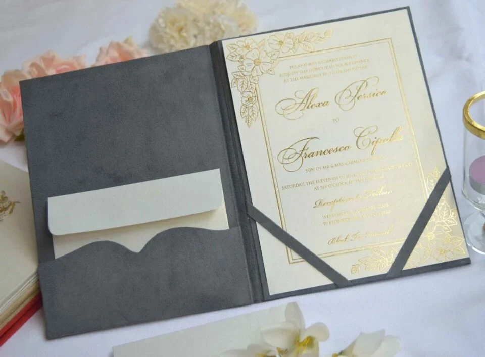An open, dark grey velvet folio wedding invitation lies flat on a white surface, surrounded by soft pink and white flowers. On the left side, a scalloped pocket holds a small cream-colored envelope. On the right side, a cream invitation card with gold foil lettering and floral designs is secured by grey corner ribbons. The card features the names "Alexa Persico" and "Francesco Cipolla" in elegant script, with a gold border framing the text.