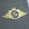 A close-up, angled shot of a dark grey velvet invitation cover. The center features a regal, gold foil-stamped crest consisting of an ornate circular frame topped with a crown and flanked by intricate leaf-like flourishes. Inside the circle, the initials are written in an elegant, interlocking gold script. The background is softly blurred, showing a hint of a white surface and a gold decorative object.