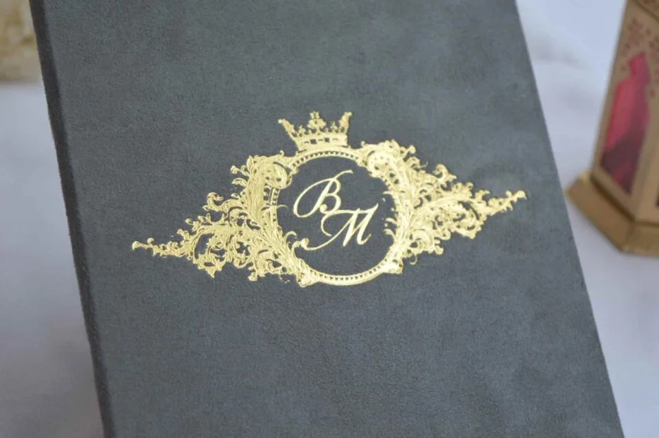 A close-up, angled shot of a dark grey velvet invitation cover. The center features a regal, gold foil-stamped crest consisting of an ornate circular frame topped with a crown and flanked by intricate leaf-like flourishes. Inside the circle, the initials are written in an elegant, interlocking gold script. The background is softly blurred, showing a hint of a white surface and a gold decorative object.