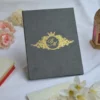 A grey velvet or suede invitation folder sits on a white surface, flanked by soft pink roses and a stack of small books. Centered on the cover is an intricate, regal gold foil crest featuring a crown, floral flourishes, and a central monogram. At the bottom of the crest, a gold ribbon banner displays the names along with the date.