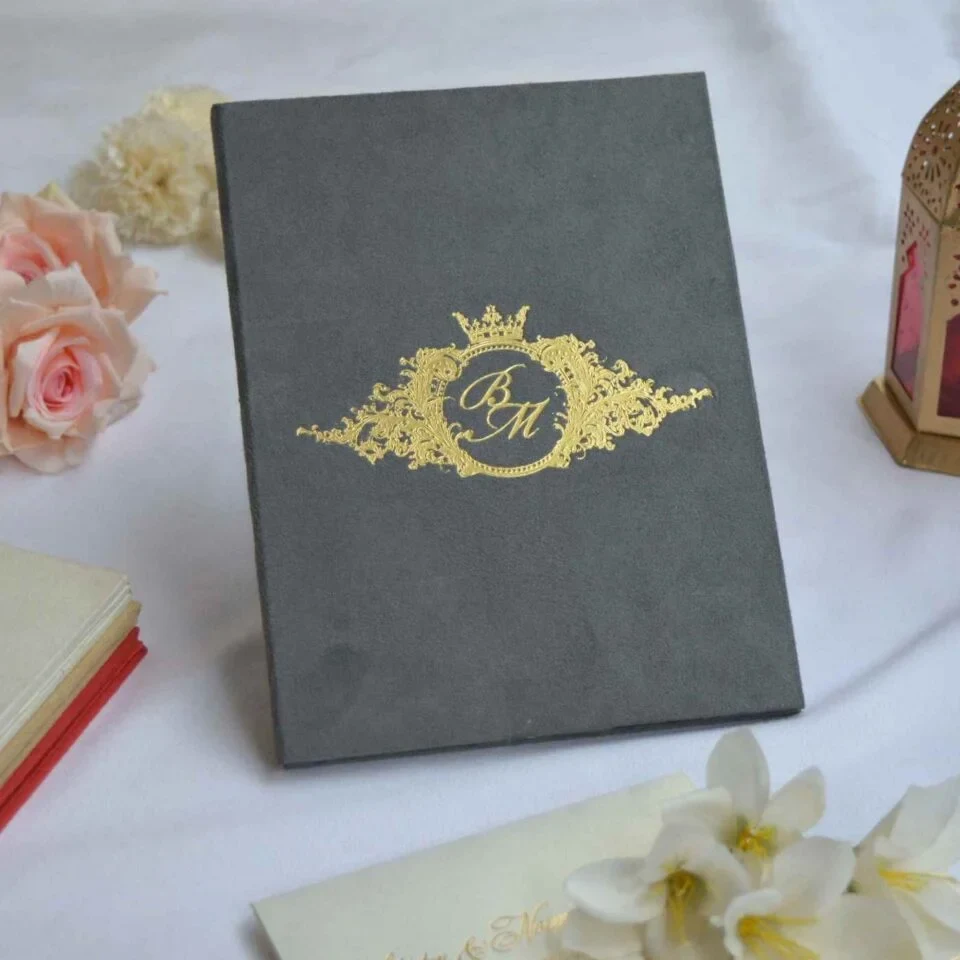 A grey velvet or suede invitation folder sits on a white surface, flanked by soft pink roses and a stack of small books. Centered on the cover is an intricate, regal gold foil crest featuring a crown, floral flourishes, and a central monogram. At the bottom of the crest, a gold ribbon banner displays the names along with the date.
