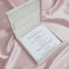 An open, **luxurious ivory wedding invitation box** resting on a backdrop of draped, shimmering **pink satin**. The interior features: * A formal **invitation card** in the base with elegant black and rose-gold typography, detailing various wedding events. * A thin, decorative rose-gold border and small flourishes at the corners of the card. * The **inner lid** contains a horizontal pocket holding a matching ivory insert or RSVP card. * The top edge of the lid reveals a glimpse of the **pearl-encrusted exterior**. * The soft, reflective folds of the pink fabric surrounding the box create a romantic and high-end aesthetic.
