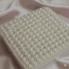 A high-angle, square shot of a **luxurious ivory-colored invitation box** resting on a draped, shimmering **pink satin fabric**. The design features: * A lid completely encrusted with a **geometric pattern of faux pearls**, arranged in precise rows and clusters. * Small **sparkling crystals** interspersed between the pearls, creating a quilted, high-shine effect. * The edges of the lid are bordered with smaller, uniform pearls for a clean finish. * The box sits at a slight angle, showcasing the smooth ivory sides and a small satin pull-tab. * The soft folds of the pink satin background provide a romantic, opulent texture that highlights the box's intricate details.