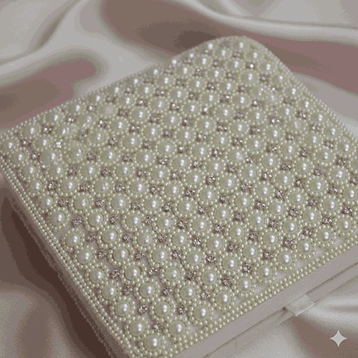 A high-angle, square shot of a **luxurious ivory-colored invitation box** resting on a draped, shimmering **pink satin fabric**. The design features: * A lid completely encrusted with a **geometric pattern of faux pearls**, arranged in precise rows and clusters. * Small **sparkling crystals** interspersed between the pearls, creating a quilted, high-shine effect. * The edges of the lid are bordered with smaller, uniform pearls for a clean finish. * The box sits at a slight angle, showcasing the smooth ivory sides and a small satin pull-tab. * The soft folds of the pink satin background provide a romantic, opulent texture that highlights the box's intricate details.