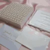 An open, **luxurious ivory wedding invitation box** resting on a backdrop of draped, shimmering **pink satin**. The interior features: * A formal **invitation card** in the base with elegant black and rose-gold typography, detailing various wedding events. * A thin, decorative rose-gold border and small flourishes at the corners of the card. * The **inner lid** contains a horizontal pocket holding a matching ivory insert or RSVP card. * The top edge of the lid reveals a glimpse of the **pearl-encrusted exterior**. * The soft, reflective folds of the pink fabric surrounding the box create a romantic and high-end aesthetic.