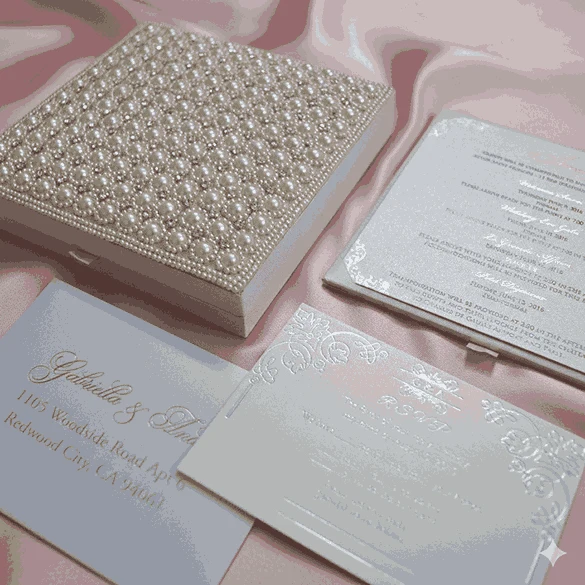 An open, **luxurious ivory wedding invitation box** resting on a backdrop of draped, shimmering **pink satin**. The interior features: * A formal **invitation card** in the base with elegant black and rose-gold typography, detailing various wedding events. * A thin, decorative rose-gold border and small flourishes at the corners of the card. * The **inner lid** contains a horizontal pocket holding a matching ivory insert or RSVP card. * The top edge of the lid reveals a glimpse of the **pearl-encrusted exterior**. * The soft, reflective folds of the pink fabric surrounding the box create a romantic and high-end aesthetic.