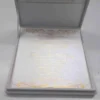 An open, **luxurious white wedding invitation box** featuring a formal stationery suite with gold accents. The interior includes: * A **white invitation card** in the base, printed with elegant **gold foil calligraphy** and framed by intricate gold scrollwork flourishes at the top and bottom. * The **inner lid** features a horizontal pocket holding a matching white card, also featuring a gold script heading. * The box is lined with a smooth, white fabric that gives it a clean and high-end appearance. * The top edge of the lid shows a hint of the **gold pearl-encrusted exterior**. * Set against a neutral, light grey background, the focus remains on the bright white textures and reflective gold detailing of the suite.