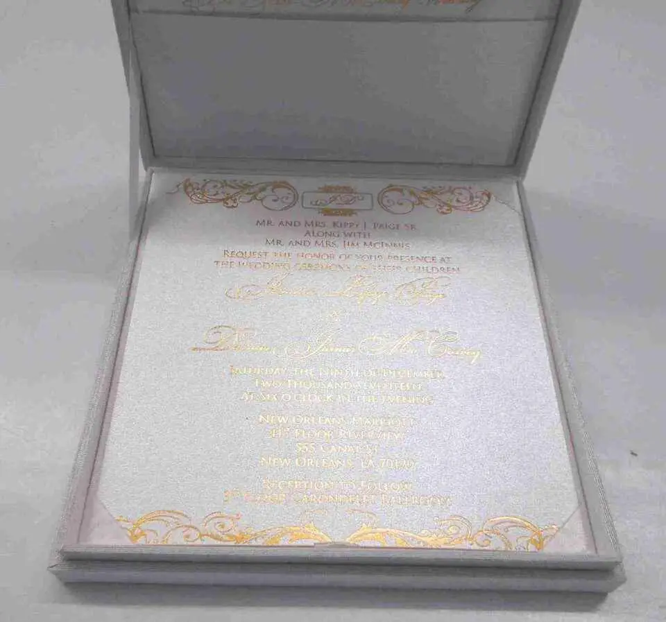 An open, **luxurious white wedding invitation box** featuring a formal stationery suite with gold accents. The interior includes: * A **white invitation card** in the base, printed with elegant **gold foil calligraphy** and framed by intricate gold scrollwork flourishes at the top and bottom. * The **inner lid** features a horizontal pocket holding a matching white card, also featuring a gold script heading. * The box is lined with a smooth, white fabric that gives it a clean and high-end appearance. * The top edge of the lid shows a hint of the **gold pearl-encrusted exterior**. * Set against a neutral, light grey background, the focus remains on the bright white textures and reflective gold detailing of the suite.