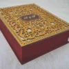 A high-angle, three-quarter view of a **luxurious burgundy invitation box** with an ornate, multi-layered lid. The box features: * A **laser-cut gold wooden overlay** on the lid, showcasing an intricate floral and scrollwork pattern. * A central **maroon plaque** featuring the custom monogram "R & F" etched in gold. * A border of **sparkling clear crystals** lining the inner edge of the gold frame. * The deep burgundy velvet sides provide a rich, tactile contrast to the metallic gold top. * The box is displayed on a neutral, textured light-colored surface, emphasizing its elegant and substantial design.