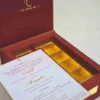 An open, **luxurious maroon wedding invitation box** featuring a multi-functional design with a built-in sweets compartment. The interior includes: * **The Invitation:** A white card printed with elegant red and gold typography for the wedding of "Rahul with Priyanka," featuring traditional Hindu motifs and "Shree Ganeshay Namah" script. * **Sweets Compartment:** To the right of the invitation, a **gold-foiled grid** provides individual sections for chocolates or traditional sweets. * **Inner Lid:** The maroon lid is accented with a thin gold border and a central gold-foiled religious inscription. * The box has a deep, textured wood-grain or fabric finish, giving it a high-end and traditional look. * The entire piece is set against a neutral, light-colored background, emphasizing the rich color palette of maroon and gold.
