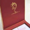 An open, **luxurious maroon wedding invitation box** featuring a multi-functional design with a built-in sweets compartment. The interior includes: * **The Invitation:** A white card printed with elegant red and gold typography for the wedding of "Rahul with Priyanka," featuring traditional Hindu motifs and "Shree Ganeshay Namah" script. * **Sweets Compartment:** To the right of the invitation, a **gold-foiled grid** provides individual sections for chocolates or traditional sweets. * **Inner Lid:** The maroon lid is accented with a thin gold border and a central gold-foiled religious inscription. * The box has a deep, textured wood-grain or fabric finish, giving it a high-end and traditional look. * The entire piece is set against a neutral, light-colored background, emphasizing the rich color palette of maroon and gold.