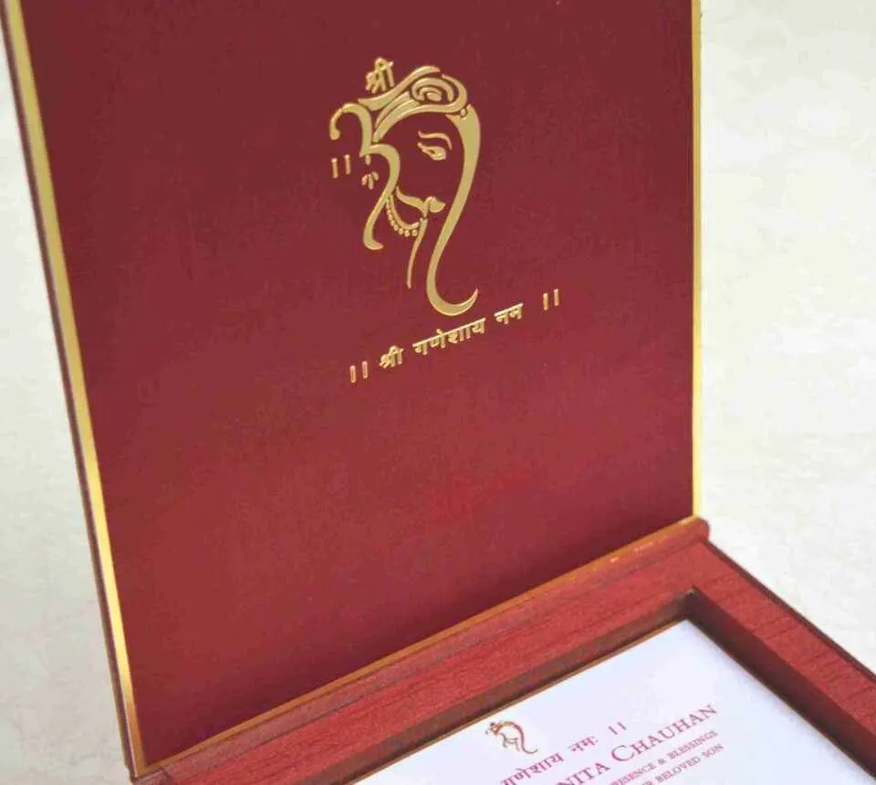 An open, **luxurious maroon wedding invitation box** featuring a multi-functional design with a built-in sweets compartment. The interior includes: * **The Invitation:** A white card printed with elegant red and gold typography for the wedding of "Rahul with Priyanka," featuring traditional Hindu motifs and "Shree Ganeshay Namah" script. * **Sweets Compartment:** To the right of the invitation, a **gold-foiled grid** provides individual sections for chocolates or traditional sweets. * **Inner Lid:** The maroon lid is accented with a thin gold border and a central gold-foiled religious inscription. * The box has a deep, textured wood-grain or fabric finish, giving it a high-end and traditional look. * The entire piece is set against a neutral, light-colored background, emphasizing the rich color palette of maroon and gold.