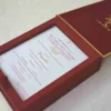 An open, **luxurious maroon wedding invitation box** featuring a multi-functional design with a built-in sweets compartment. The interior includes: * **The Invitation:** A white card printed with elegant red and gold typography for the wedding of "Rahul with Priyanka," featuring traditional Hindu motifs and "Shree Ganeshay Namah" script. * **Sweets Compartment:** To the right of the invitation, a **gold-foiled grid** provides individual sections for chocolates or traditional sweets. * **Inner Lid:** The maroon lid is accented with a thin gold border and a central gold-foiled religious inscription. * The box has a deep, textured wood-grain or fabric finish, giving it a high-end and traditional look. * The entire piece is set against a neutral, light-colored background, emphasizing the rich color palette of maroon and gold.