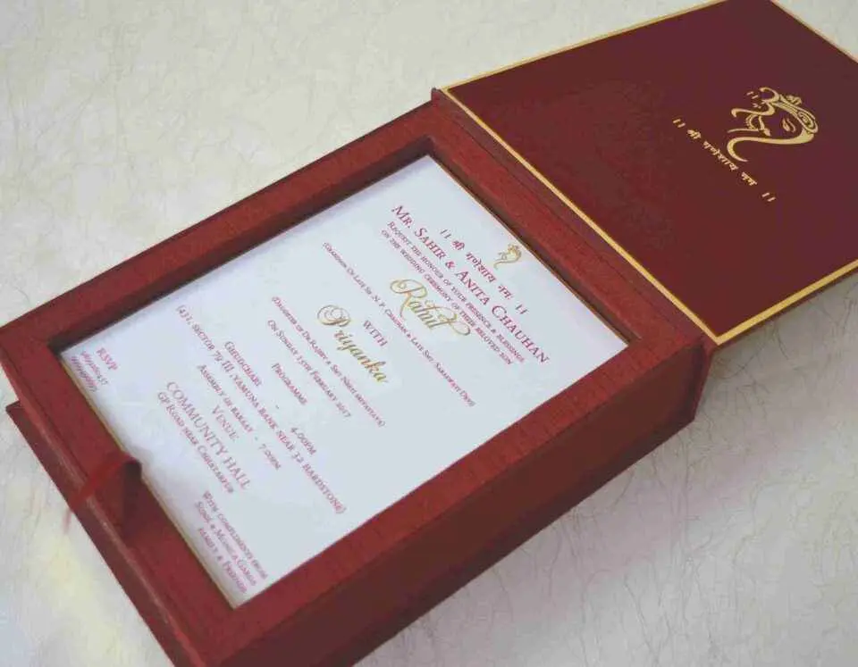 An open, **luxurious maroon wedding invitation box** featuring a multi-functional design with a built-in sweets compartment. The interior includes: * **The Invitation:** A white card printed with elegant red and gold typography for the wedding of "Rahul with Priyanka," featuring traditional Hindu motifs and "Shree Ganeshay Namah" script. * **Sweets Compartment:** To the right of the invitation, a **gold-foiled grid** provides individual sections for chocolates or traditional sweets. * **Inner Lid:** The maroon lid is accented with a thin gold border and a central gold-foiled religious inscription. * The box has a deep, textured wood-grain or fabric finish, giving it a high-end and traditional look. * The entire piece is set against a neutral, light-colored background, emphasizing the rich color palette of maroon and gold.