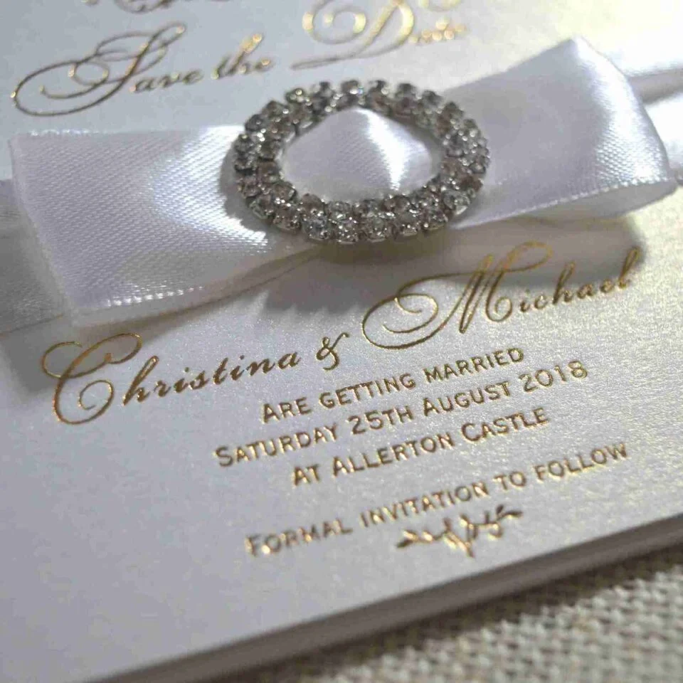 Ivory silk save the date card, adorned with a stunning rhinestone brooch on satin ribbon and printed with gold foil.